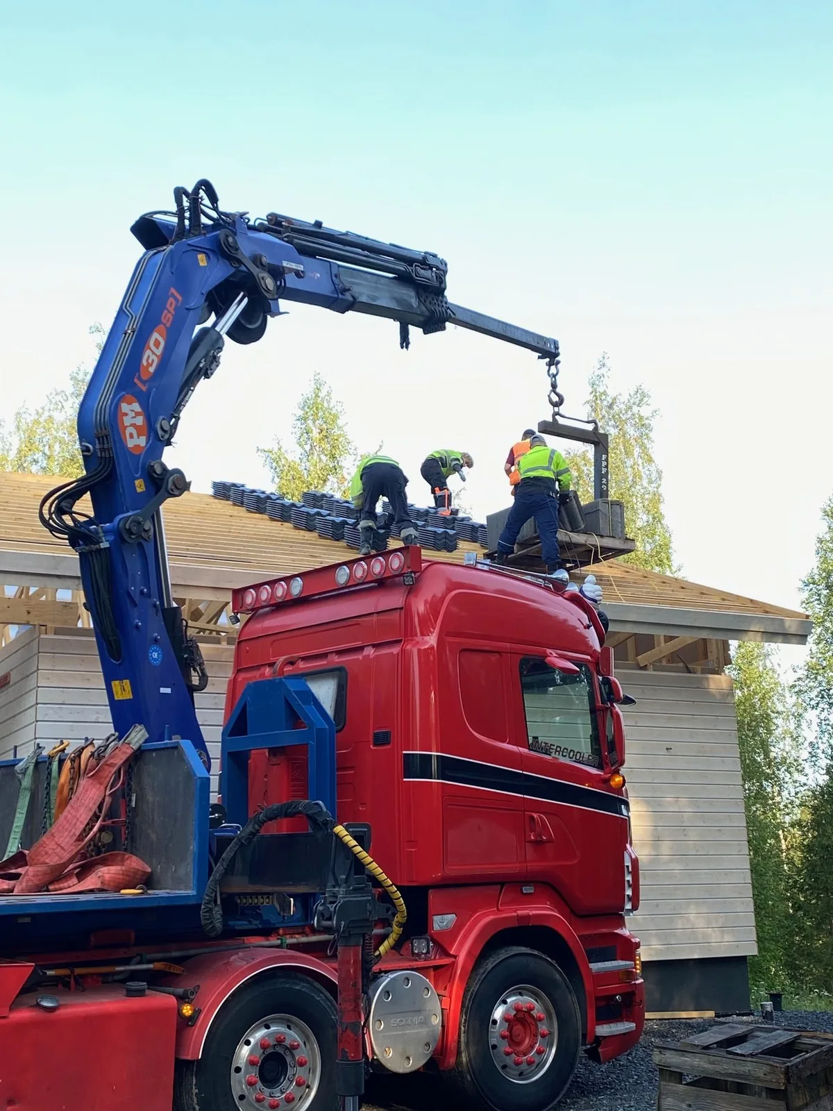 Nosturi nostaa kattotiilipaletin katolle, kolme asentajaa vastaanottamassa.